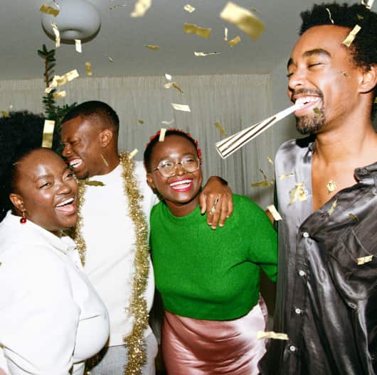 Four people laughing with their arms around one another's shoulders. One has a party popper in his mouth, another is wearing a gold streamer around his shoulders, and there is gold confetti in the air.