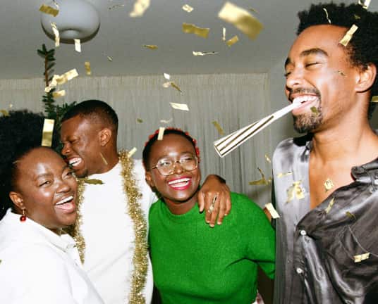 Four people laughing with their arms around one another's shoulders. One has a party popper in his mouth, another is wearing a gold streamer around his shoulders, and there is gold confetti in the air.
