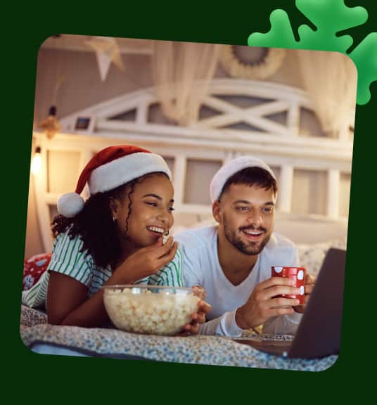 A woman and man wearing santa hats in bed looking at a laptop linking to holiday movie DVDs page