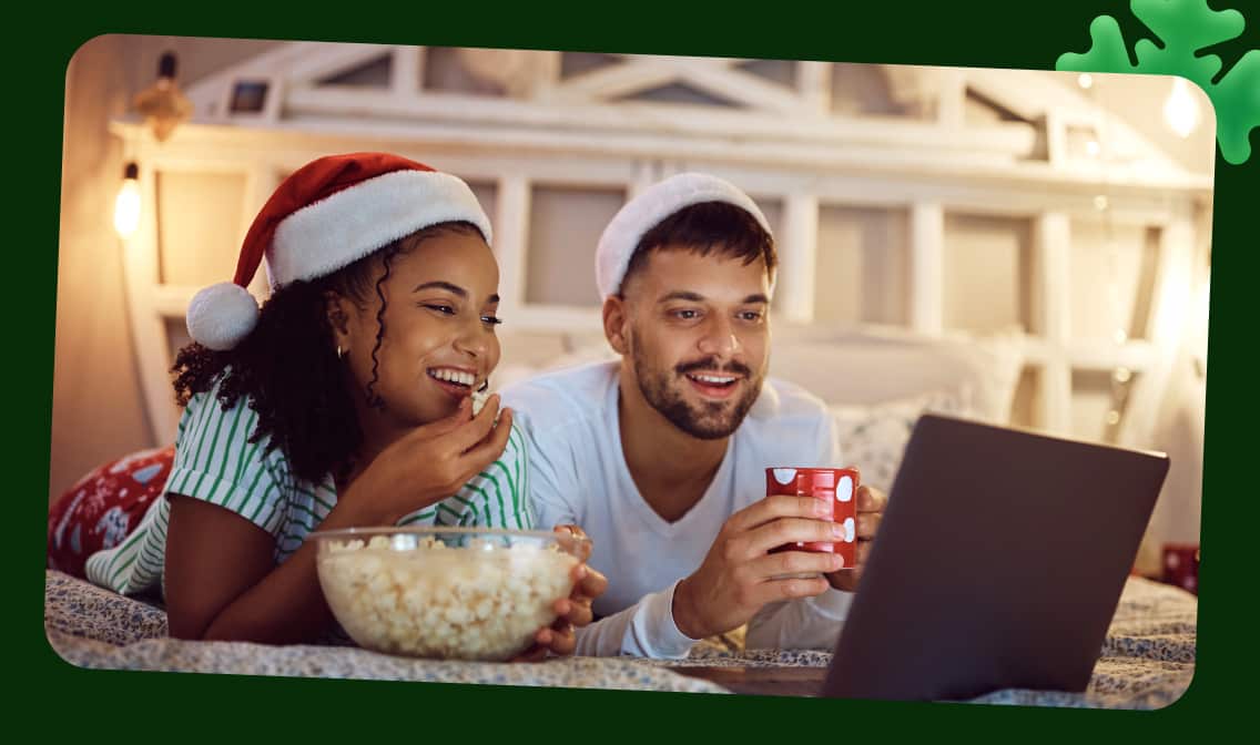 A woman and man wearing santa hats in bed looking at a laptop linking to holiday movie DVDs page
