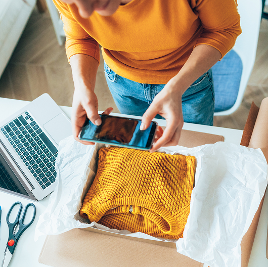 A woman packing up and taking a picture of an orange sweater, linking to the selling page.
