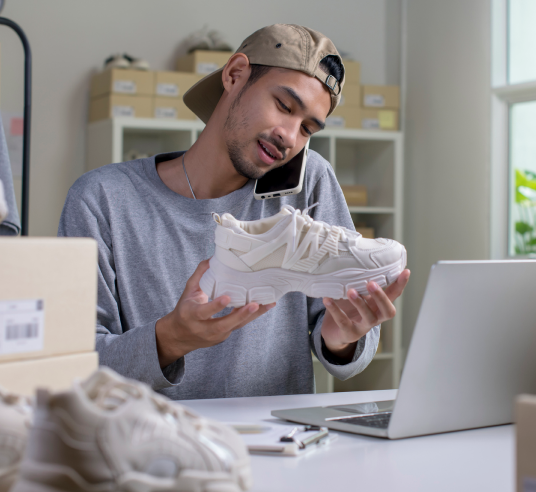 A man in a baseball cap examines a white athletic shoe. Links to Selling page.