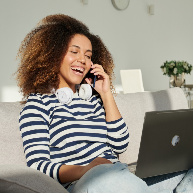 A smiling woman on a cell phone sits on a sofa while working on her laptop. Image links to eBay customer service page.