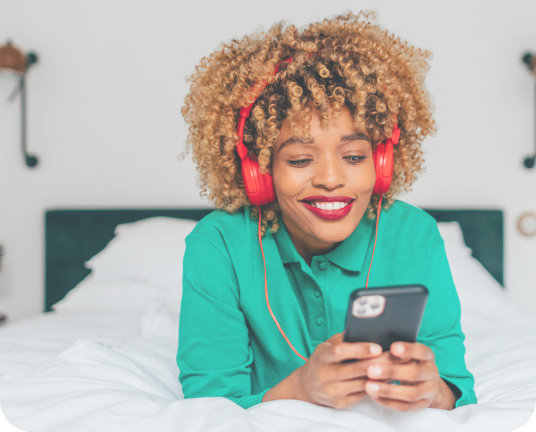 A young woman wearing red over the ear headphones lounging on a bed and looking at her smartphone.